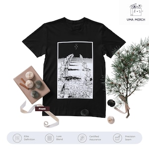 May include: Black t-shirt featuring a black and white graphic print. The graphic depicts a line of figures in a desolate landscape. The shirt is displayed with decorative paper ornaments and a small evergreen branch.