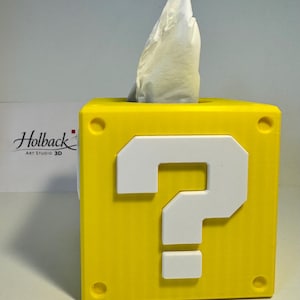 May include: A bright yellow square tissue box cover shaped like a question mark block. The front face features a large, raised white question mark symbol. A white tissue is protruding from the top opening of the cube-shaped container.