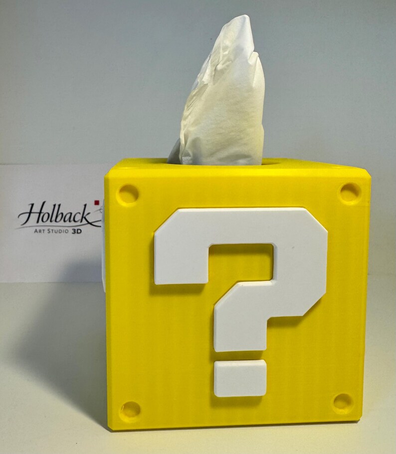 May include: A bright yellow square tissue box cover shaped like a question mark block. The front face features a large, raised white question mark symbol. A white tissue is protruding from the top opening of the cube-shaped container.
