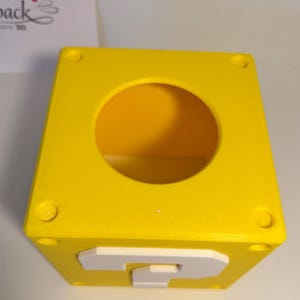May include: A bright yellow cube-shaped container with a large circular opening on the top surface. The front face features a raised white question mark symbol. The top corners of the cube have small circular indentations, creating a block design.