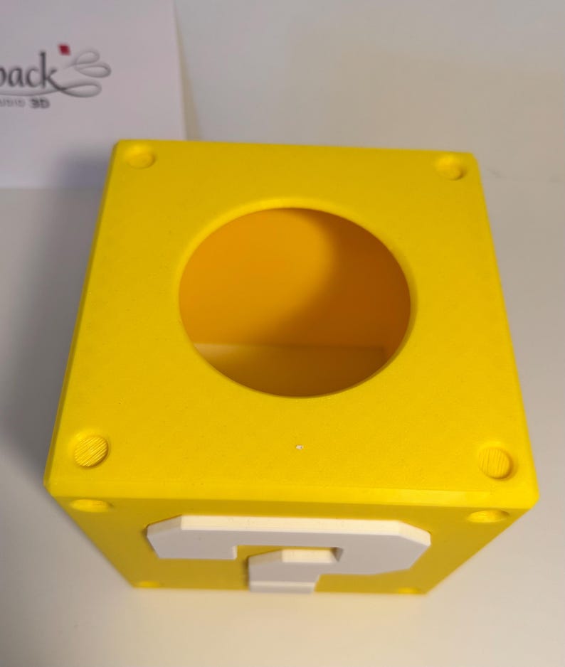 May include: A bright yellow cube-shaped container with a large circular opening on the top surface. The front face features a raised white question mark symbol. The top corners of the cube have small circular indentations, creating a block design.