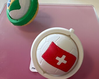 May include: Two spherical objects featuring soccer ball patterns encased in circular frames. One object is white with a red Swiss flag emblem, while the other is green and yellow with a blue and yellow emblem. Both items have a textured finish.