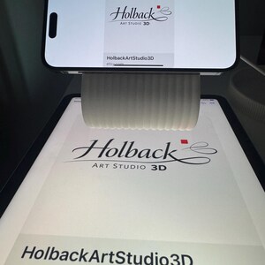 May include: A white, ribbed, cylindrical phone stand holding a smartphone horizontally. Both the smartphone and the tablet underneath display the logo for Holback Art Studio 3D, featuring the text Holback Art Studio 3D and the website etsy.com.