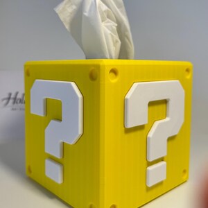 May include: A bright yellow square tissue box cover featuring a raised white question mark on each visible side. The cube-shaped container has small circular indentations at each corner and a white tissue protruding from the top opening.