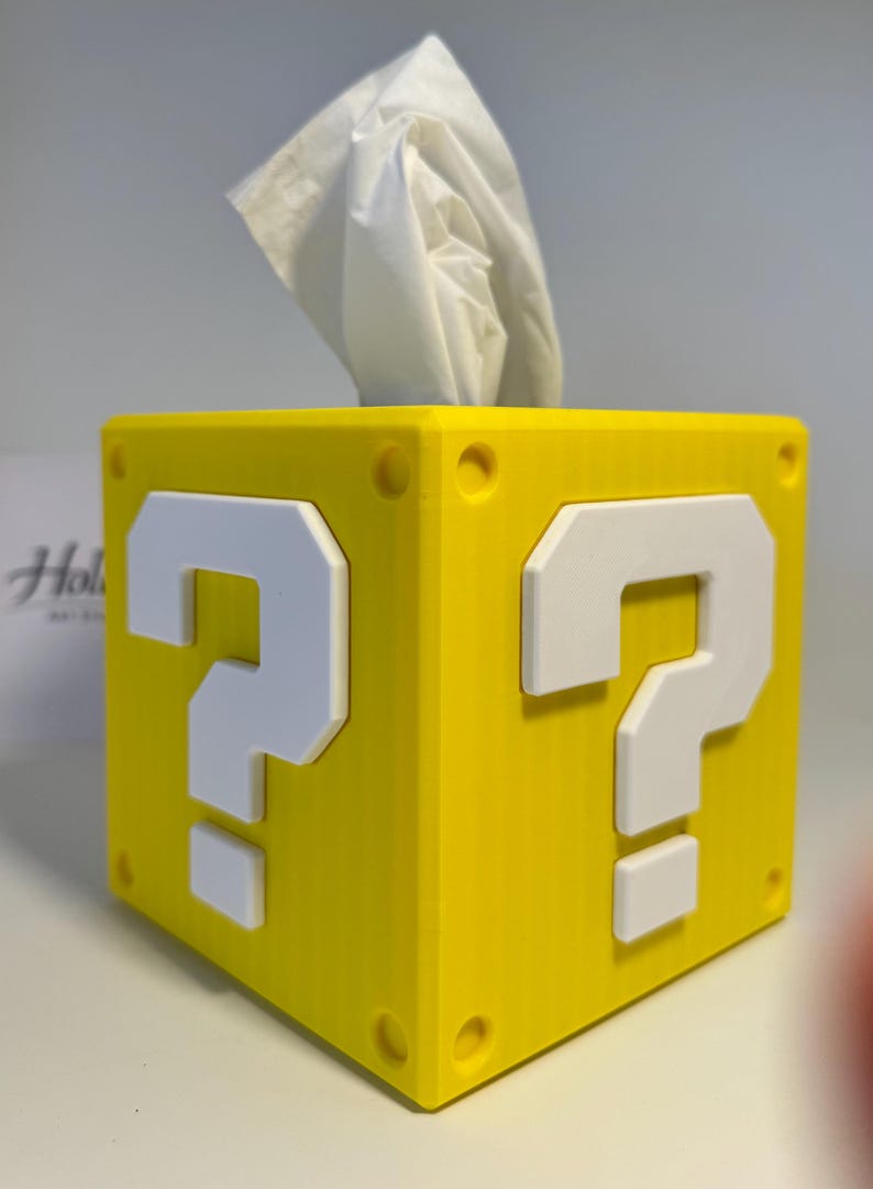 May include: A bright yellow square tissue box cover featuring a raised white question mark on each visible side. The cube-shaped container has small circular indentations at each corner and a white tissue protruding from the top opening.