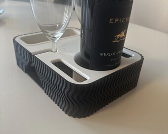May include: A black and white wine bottle and glass holder. The holder has a wavy edge design and holds a wine bottle and two wine glasses. The wine bottle has a black label with gold text. The holder is on a white surface.
