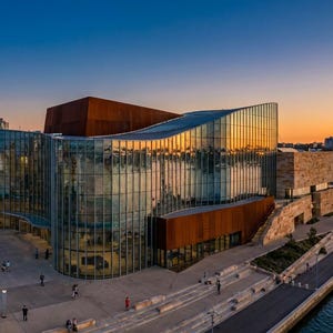 May include: Panoramic view of a modern architectural complex with glass and rust-colored metal facades. The building reflects the sunset, with a city skyline in the background. A waterfront promenade is visible.