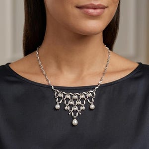 Puede incluir: Un collar de plata con un diseño delicado que presenta múltiples perlas pequeñas y un colgante de perla más grande en forma de lágrima. El collar se usa sobre un fondo negro.