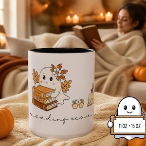 May include: A white ceramic mug with a black interior featuring a cartoon ghost holding a stack of books, decorated with autumn leaves and flowers. The text "reading season" is printed below the illustration. A small ghost icon displays 11 oz + 15 oz.