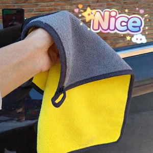 Puede incluir: Un paño de limpieza de microfibra amarillo y gris con un borde gris oscuro. El paño se sostiene en alto y la palabra "Nice" es visible en una fuente decorativa en la esquina superior derecha.