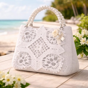 Puede incluir: Un bolso blanco de ganchillo con un asa de perlas y un adorno floral decorativo. El bolso presenta intrincados detalles de encaje. El bolso está sobre un fondo de playa, mostrando un diseño delicado y elegante.