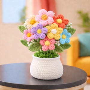 May include: A handmade crochet flower arrangement in a white pot. The colourful flowers include pink, orange, yellow, blue, and purple petals with green leaves. The pot is sitting on a dark surface.