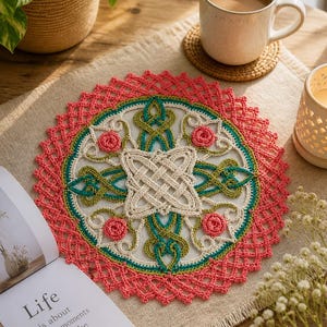May include: A handmade, circular crochet doily with a detailed Celtic knot design. The doily features a cream-colored center with green, pink, and cream accents. The outer edge is a vibrant coral color. A cup of coffee and a book are also visible.