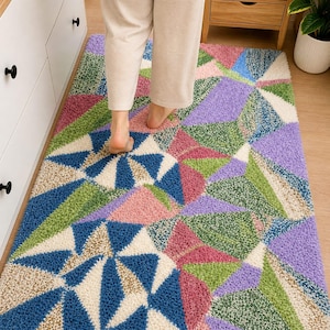 May include: A colorful geometric rug with a mix of blue, green, pink, purple, and cream triangular shapes. The rug has a shaggy texture and is placed on a wooden floor. The rug is rectangular and appears to be in a bathroom or bedroom.