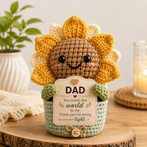May include: A crocheted sunflower figurine with a smiling face, yellow petals, and a brown center. It holds a sign that reads "DAD" and a message. The figurine sits in a green pot with a wooden button.