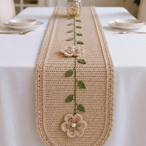 May include: A beige crochet table runner featuring a central vine pattern with green leaves and two dimensional beige flowers. Each flower has a white pearl bead at its center. The runner has a rounded end with a beige tassel and a pearl bead.