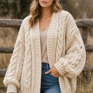 Könnte beinhalten: Cremefarbene Strickjacke mit passender Mütze. Der offene Cardigan hat ein grobes Strickmuster, einen V-Ausschnitt und lange Ärmel. Das Model trägt eine blaue Jeans und ein beiges Oberteil. Die Länge der Strickjacke beträgt ca. 76 cm.