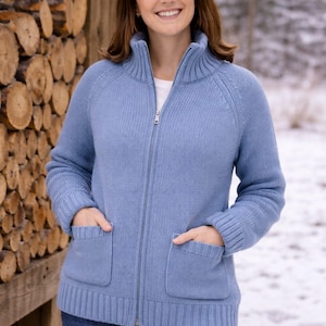 Könnte beinhalten: Hellblauer Strick-Reißverschluss-Cardigan mit hohem Kragen und zwei Vordertaschen. Der Cardigan wird mit einer blauen Jeans getragen. Der Hintergrund zeigt eine verschneite Landschaft und einen Holzstapel. Das Material ist gestrickt.