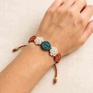 May include: A bracelet featuring a series of crocheted elements including two cream-colored flowers, a central dark teal leaf-shaped piece, and several rust-colored leaf-shaped accents, all connected by a rust-colored cord with small gold-toned beads.