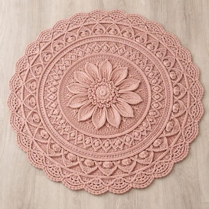 May include: A round, dusty rose-colored crochet rug with a floral design. The rug features a large flower in the center, surrounded by intricate patterns and a scalloped edge. The rug is made of soft yarn and is suitable for home decor.