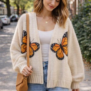 Puede incluir: Cárdigan color crema con dos grandes aplicaciones de mariposas en naranja, negro y blanco. El cárdigan abierto tiene mangas largas y se usa con una camiseta blanca y vaqueros azules. Un bolso marrón cuelga del hombro.