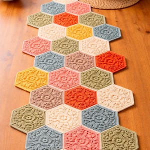 May include: A colorful, handmade crochet table runner. The runner is composed of hexagonal shapes in various colors, including cream, orange, yellow, pink, and green. Each hexagon features a floral design in the center. The runner is placed on a wooden table.