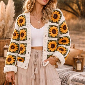 Op de afbeelding: Crèmekleurig gehaakt vest met vierkante zonnebloemmotieven in geel, bruin en groen. Het vest heeft lange mouwen en een knoopsluiting. De outfit omvat een witte top, een beige rok en een bruine hoed. Het thema is bohemien.