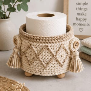 May include: A cream-colored, crocheted toilet paper holder with a diamond pattern. The holder has a tan border, wooden feet, and decorative tassels. A roll of white toilet paper sits inside. The image includes a framed sign that reads "simple things make happy moments."