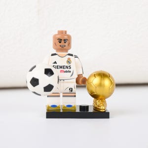 May include: A Lego figurine of a soccer player in a white jersey with black accents, holding a soccer ball and standing next to a golden trophy. The jersey has the number 9 and the Siemens Mobile logo.