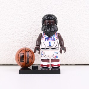 Puede incluir: Figura Lego de un jugador de baloncesto con una camiseta blanca con "PHILA" y el número "1". La figura tiene barba negra y sostiene una pelota de baloncesto marrón con "NBA" y "SPALDING" impresos.