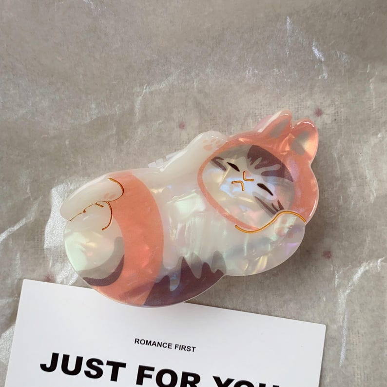 May include: A cat-shaped hair clip in shades of pink, white, and brown, with a pearlescent finish. The clip features a sleeping cat design with gold-colored accents. The text "ROMANCE FIRST JUST FOR YOU" is visible on a white card.