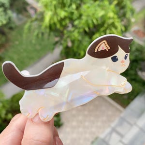 May include: A white and brown cat-shaped hair clip with a pearlescent finish. The cat has brown ears and a tail, with gold-colored accents. The clip is designed to hold hair in place and adds a playful touch to any hairstyle.