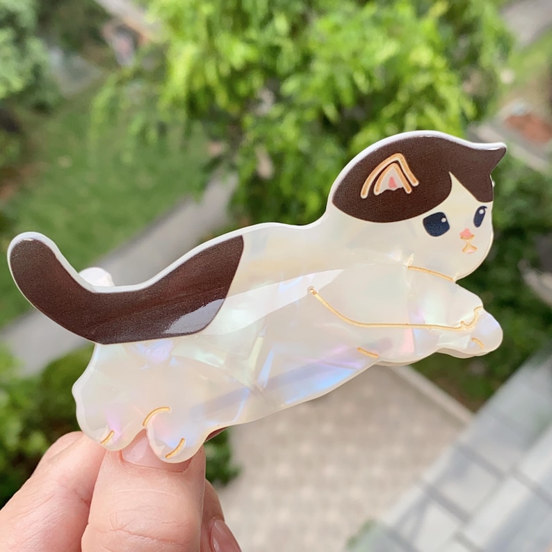 May include: A white and brown cat-shaped hair clip with a pearlescent finish. The cat has brown ears and a tail, with gold-colored accents. The clip is designed to hold hair in place and adds a playful touch to any hairstyle.