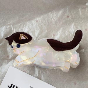 May include: A cat-shaped hair clip in iridescent white with brown markings. The clip features a playful design with gold accents and blue eyes. The cat is lying down, with its tail curved upwards. The clip is made of a shiny material.