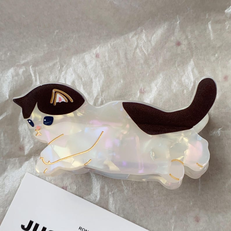 May include: A cat-shaped hair clip in iridescent white with brown markings. The clip features a playful design with gold accents and blue eyes. The cat is lying down, with its tail curved upwards. The clip is made of a shiny material.