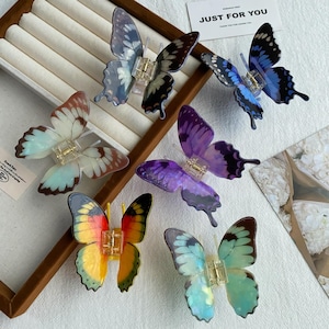 May include: Six butterfly-shaped hair clips in various colors including blue, purple, yellow, orange, and translucent white with brown edges. Each clip features a central metal spring mechanism and translucent wings with detailed patterns.