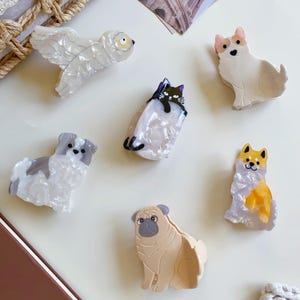 May include: A collection of animal-shaped hair clips. The clips include an owl, a cat, a corgi, a shiba inu, a pug, and a Shih Tzu. The clips are made of a pearlescent material with various colors, including white, gray, and tan.