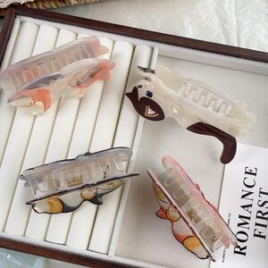 May include: Four cat-shaped hair claw clips in a variety of colors. The clips are made of translucent plastic and feature painted cat designs in shades of brown, orange, and gray. The clips are arranged on a white surface.