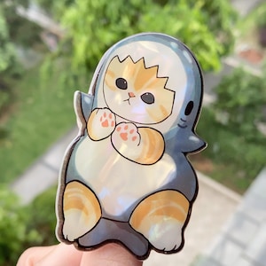 May include: A cute sticker featuring a cat in a shark costume. The cat has orange and white fur, pink paw pads, and is inside a blue and white shark costume. The sticker has a pearlescent finish and is held up by a finger.