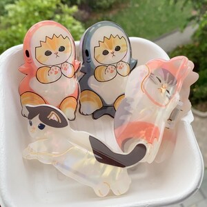 May include: Four cat-themed hair clips in a white container. Two clips feature orange and white cats in shark costumes, one in pink and the other in blue. The other two clips are cat-shaped, one white with black and brown markings, and the other with pink and orange stripes.