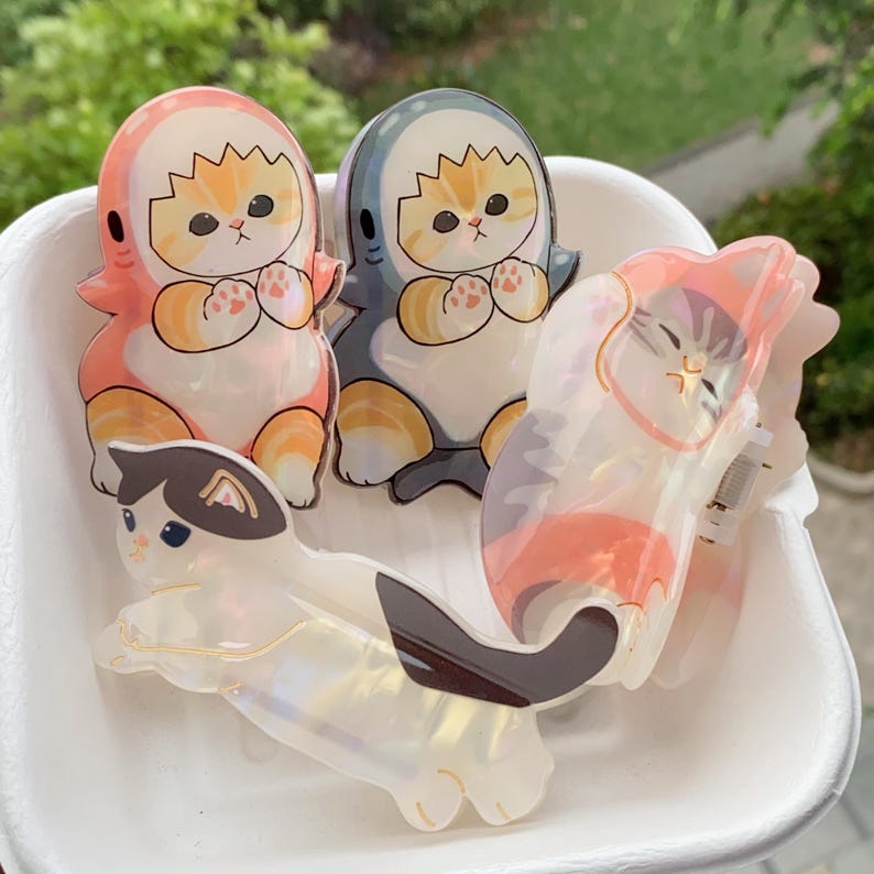 May include: Four cat-themed hair clips in a white container. Two clips feature orange and white cats in shark costumes, one in pink and the other in blue. The other two clips are cat-shaped, one white with black and brown markings, and the other with pink and orange stripes.