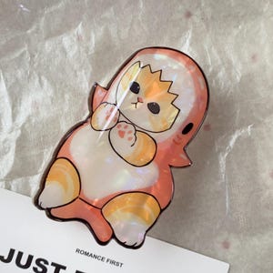 May include: A whimsical, iridescent cat-shaped pin, dressed in a shark costume. The pin features orange, white, and pink hues, with a glossy finish. The text "ROMANCE FIRST" is visible at the bottom.