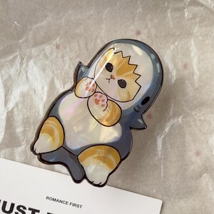 May include: A whimsical cat-shaped pin, designed to resemble a shark. The pin features a calico cat with orange and white fur, wearing a blue and white shark costume. The pin has a pearlescent finish and is set against a white background.