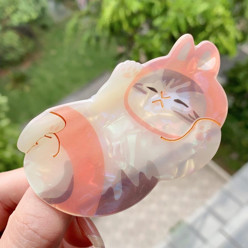 May include: A pearlescent cat-shaped hair clip in a sleeping pose. The clip features a pink and orange hooded design with a brown and white cat face. The cat is lying down with its eyes closed, and the clip has a gold-colored outline.