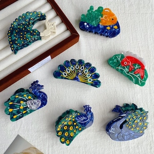 May include: A collection of six acetate hair claw clips featuring animal designs. Included are four peacock-shaped clips in shades of blue and green with yellow accents, and two fish-shaped clips, one in orange and blue and one in red and green.