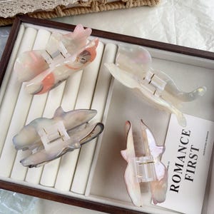May include: Four iridescent hair claw clips in a display case. The clips are shaped like butterflies and feature a pearlescent finish with shades of pink, white, and gray. Each clip has a metal spring mechanism for securing hair.