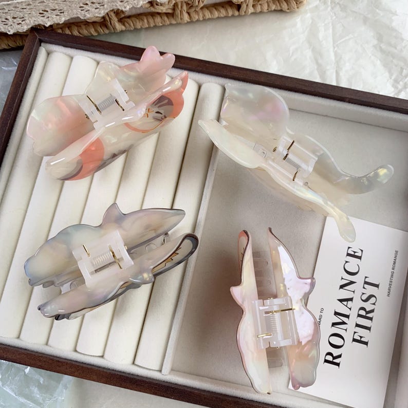 May include: Four iridescent hair claw clips in a display case. The clips are shaped like butterflies and feature a pearlescent finish with shades of pink, white, and gray. Each clip has a metal spring mechanism for securing hair.
