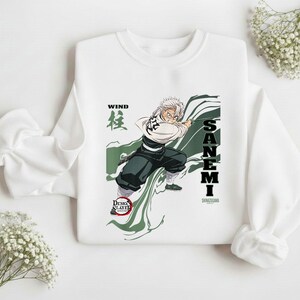 Puede incluir: Sudadera blanca con un gr&aacute;fico de un personaje de anime, Sanemi, en acci&oacute;n con el texto "WIND" y "SANEMI". El dise&ntilde;o incluye acentos verdes y negros, y el logo "Demon Slayer".