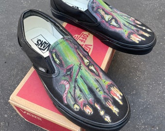 May include: A pair of black Vans slip-on shoes with a hand-painted design of a green zombie hand. The shoes have the Vans logo on the side and the words "Off The Wall" on the back.