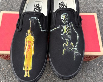 May include: A pair of black Vans slip-on shoes with a graphic print. One shoe features a skeleton holding a woman by the neck, while the other shoe features a woman hanging from a rope. The shoes have the Vans logo on the back.